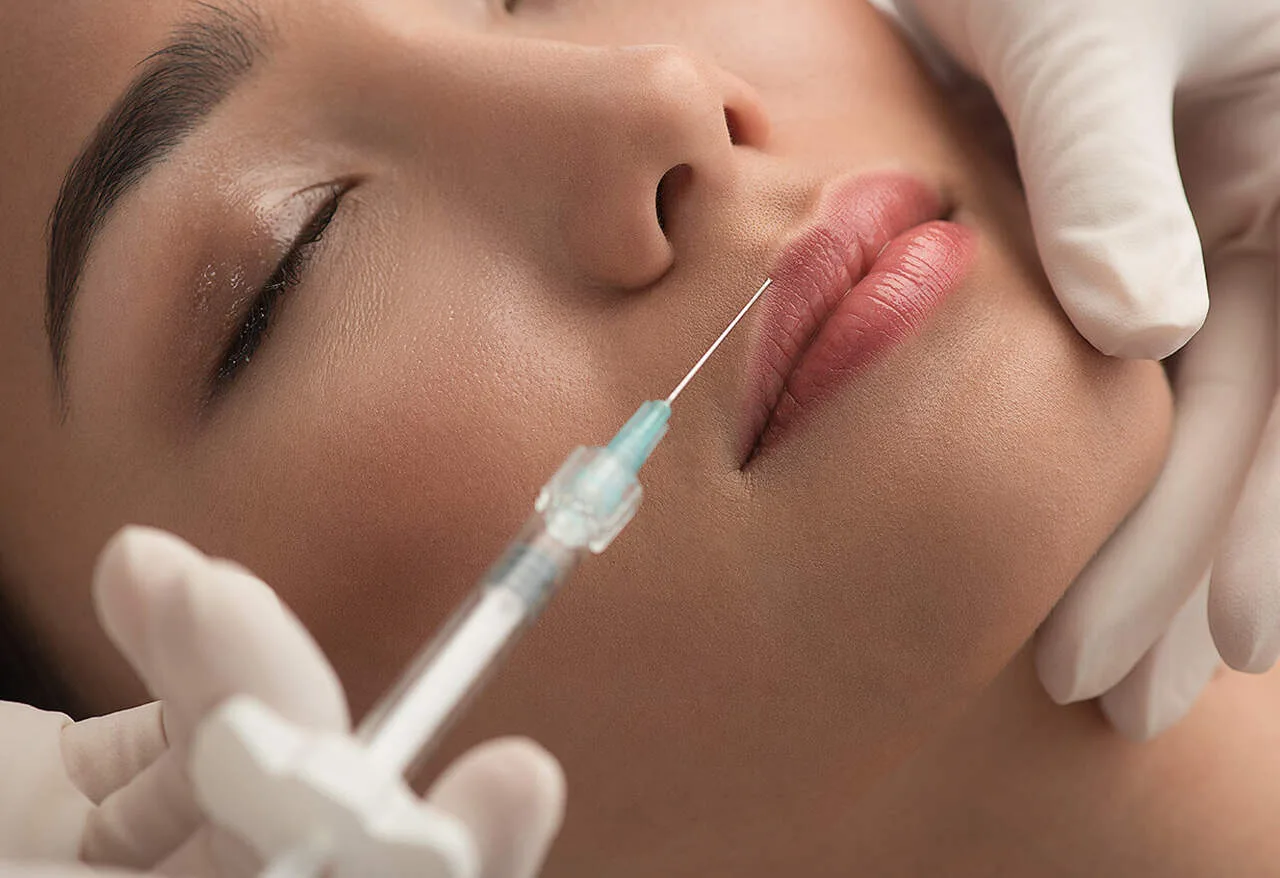 profissional com seringa fazendo harmoniza&ccedil;&atilde;o facial com mulher de olhos fechados.